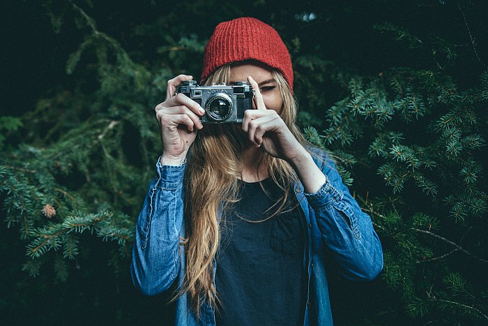 Photographer with a red hat, standing in front of a fir tree, taking a picture with her retro camera.